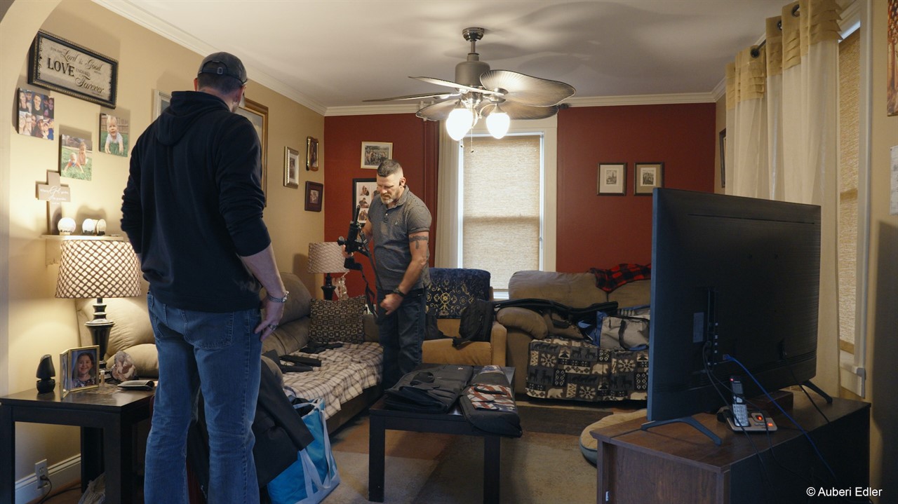 Two men stand in a living room, one holding a rifl