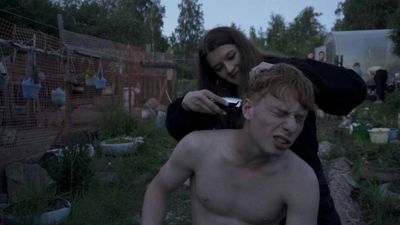 A teen girl shaves a teen boy's head
